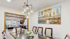 A dining area with a long table, chairs, a chandelier, and a painting of a landscape. - Lakeside Chalet with Hot Tub, Fire Pit & Game Room (Blakeslee)