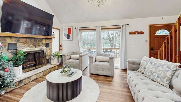 A spacious living room with a fireplace, comfortable seating, a large TV, and a view through the windows. - Lakeside Chalet with Hot Tub, Fire Pit & Game Room (Blakeslee)