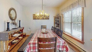 A dining room with a long table, chairs, a chandelier, a mirror, and a window with blinds. - Cozy Home Near Waterpark - Hot Tub and Deck (Tannersville)