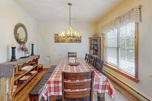 A dining room with a long table, chairs, a chandelier, a mirror, and a window with blinds. - Cozy Home Near Waterpark - Hot Tub and Deck (Tannersville)