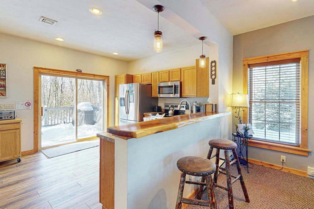 A spacious kitchen with wooden cabinets, a central island with stools, a large window, and a sliding glass door leading to a deck.