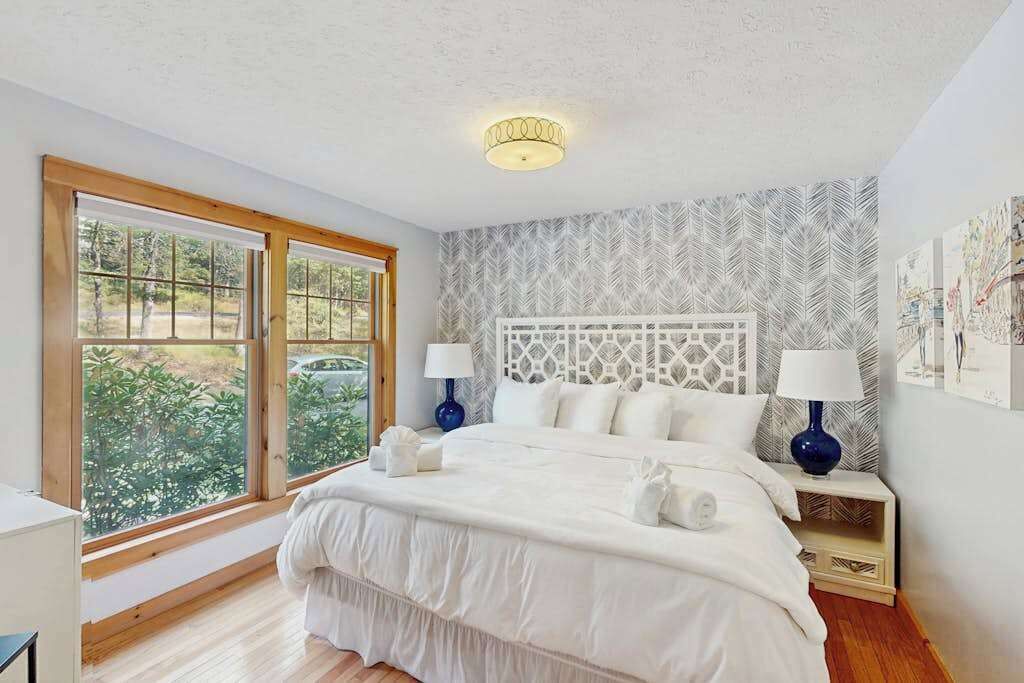 Une chambre avec un grand lit, deux tables de chevet avec des lampes, un luminaire au plafond, une fenêtre donnant sur un paysage agréable et un mur décoré d’un motif.