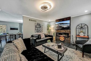 A modern living room with a fireplace, a dining area, and a kitchen in the background. - Spacious House with Hot Tub Game Room and Firepit (Long Pond)