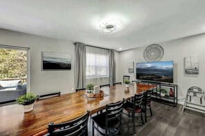 A dining area with a wooden table, chairs, a large window with curtains, a wall-mounted TV, and a view of the outdoors. - Long Pond Home with Hot Tub, Billiards & Poker (Long Pond)