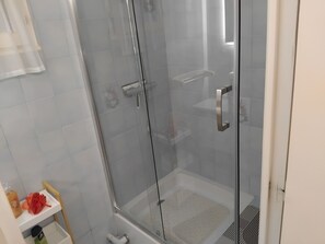 A glass-enclosed shower with a handheld showerhead, a white tiled wall, and a small shelf with toiletries.