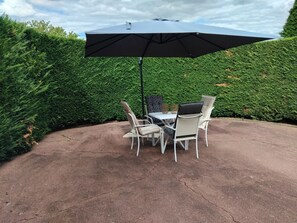 A patio with a table and chairs under a large umbrella, surrounded by a hedge.