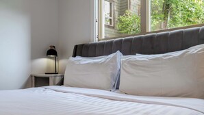 Una cama con sábanas blancas, una lámpara de noche y una ventana con vistas a zonas verdes.