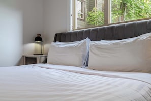 Una cama con sábanas blancas, una lámpara de noche y una ventana con vistas a zonas verdes.