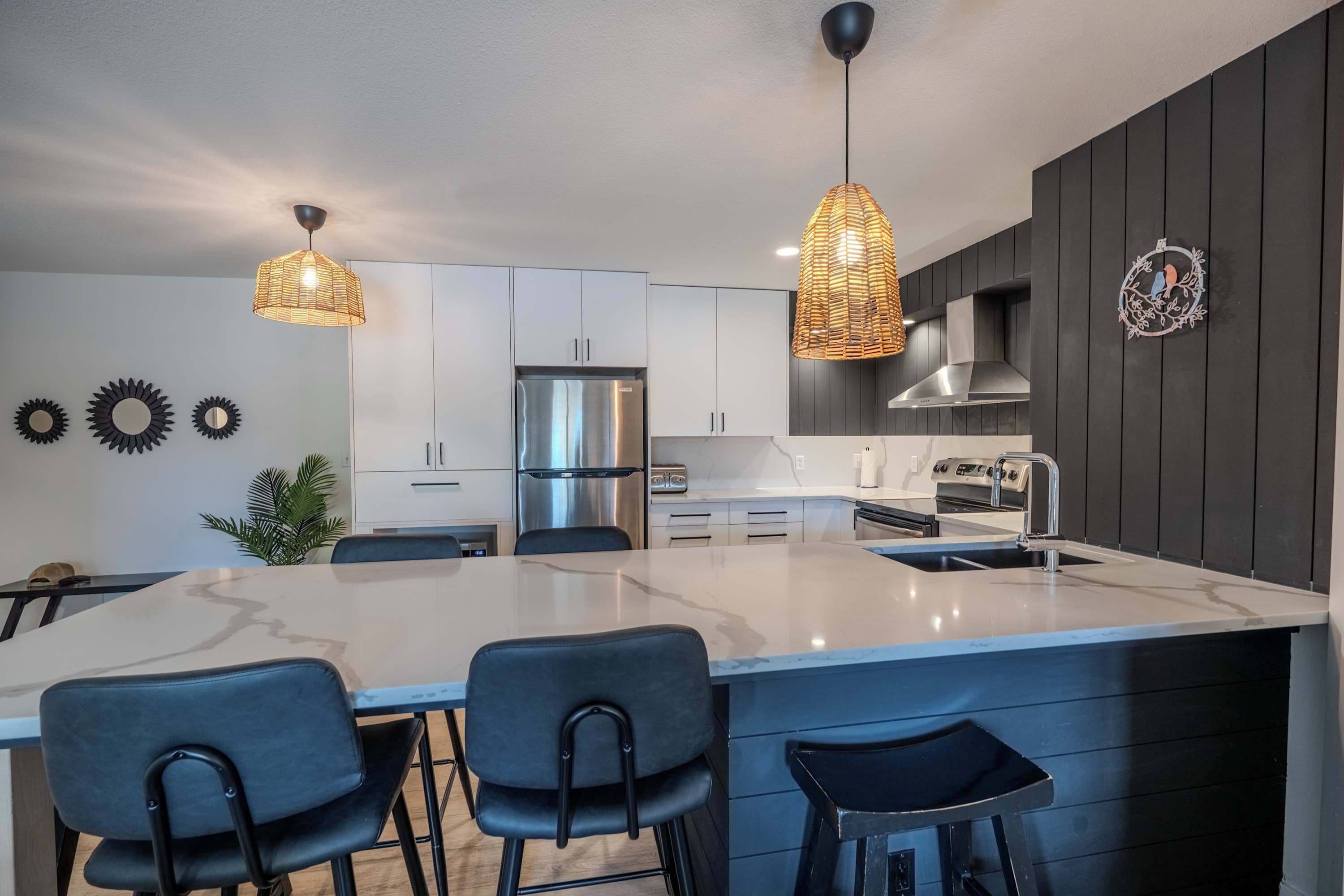 Une cuisine moderne dotée d’un îlot central, de luminaires suspendus, d’appareils électroménagers en acier inoxydable et de meubles de rangement aux couleurs sombres.