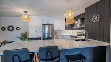 Une cuisine moderne dotée d’un îlot central, de luminaires suspendus, d’appareils électroménagers en acier inoxydable et de meubles de rangement aux couleurs sombres.