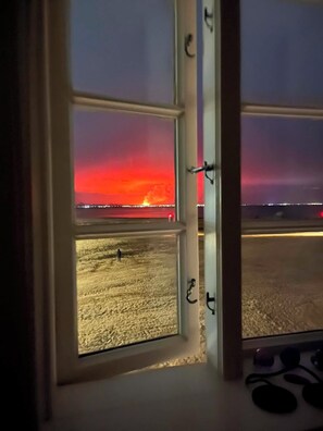A window with a view of a beach at sunset, a pier in the distance, and a person walking on the sand.