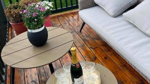 Una terraza de madera con un sofá de mimbre, una mesita con un jarrón de flores y una botella de champán.