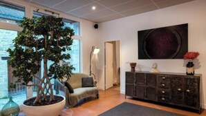 A modern living room with a large potted tree, a sofa, a floor lamp, a dark console table with decorative items, and a large abstract painting on the wall.