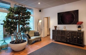 Un salon moderne avec un grand arbre en pot, un canapé, une lampe sur pied, une console sombre agrémentée d’objets décoratifs et un grand tableau abstrait accroché au mur.