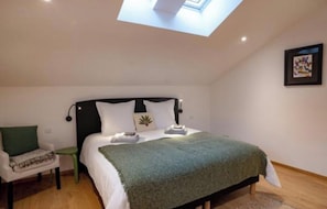 Une chambre avec un puits de lumière, un lit recouvert d’une couverture verte, une table de chevet et une chaise.