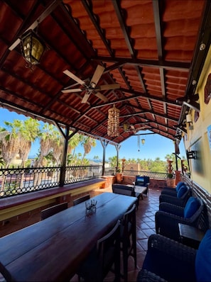 An outdoor patio with wooden tables and chairs, a hanging light fixture, and a view of palm trees. - Casa Valentina - Tuna Suite - Right Downtown and Beach (Loreto)