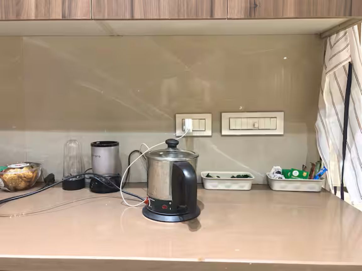 A kitchen counter with a kettle, a blender, and a food processor. There are two wall-mounted light switches and a white cabinet above the counter.