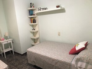 Une chambre avec un lit, une table de chevet, une étagère avec des livres et des figurines, et une étagère murale sur laquelle est accroché un panneau indiquant « Chez soi ».