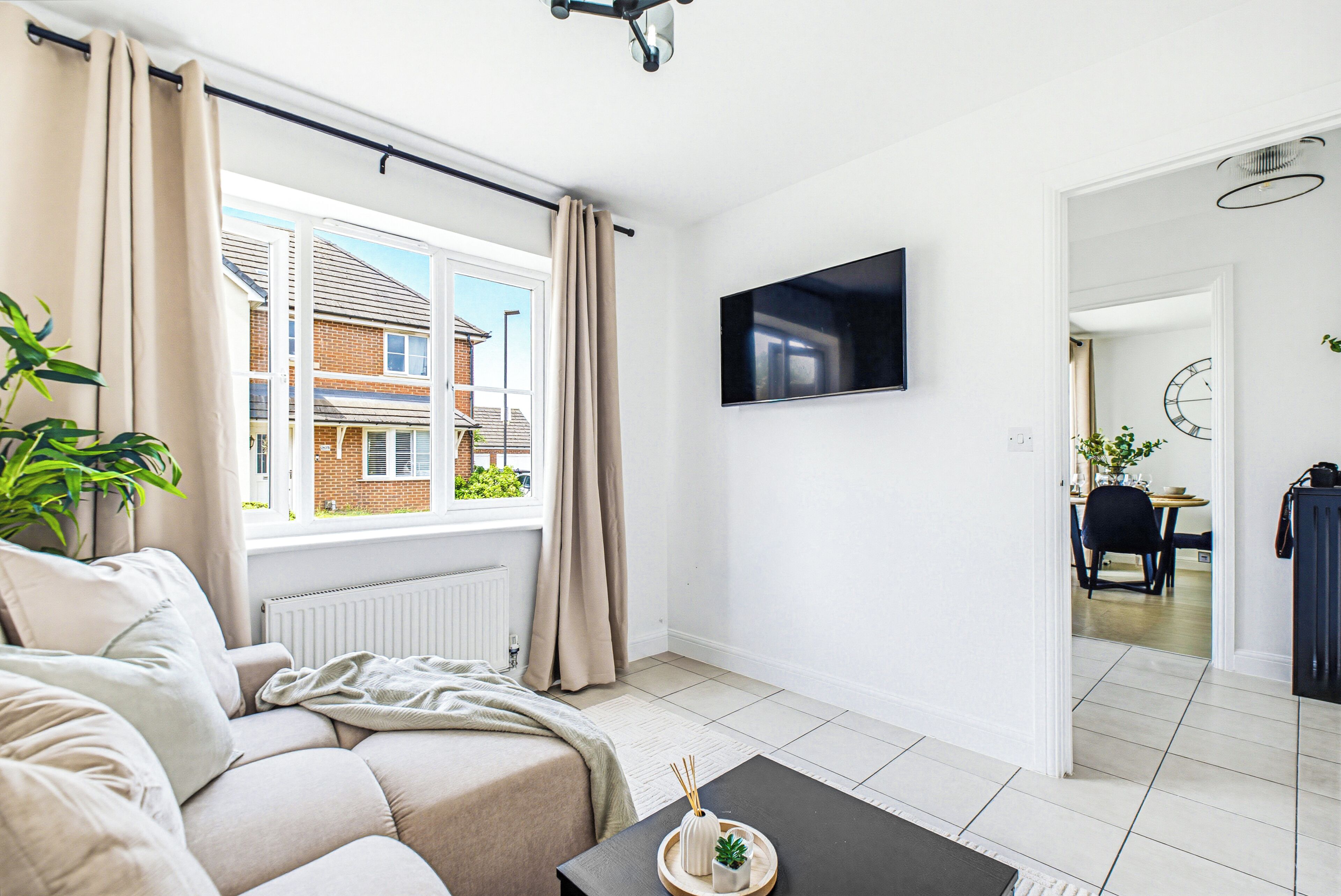Una sala de estar con un sofá, una mesa de centro con una planta, un televisor en la pared y vista a una casa a través de una ventana.