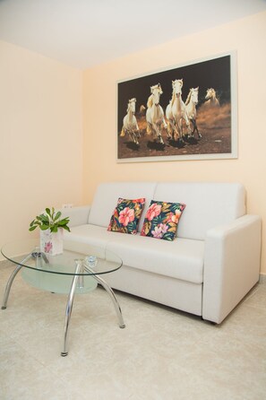 Un canapé blanc agrémenté de coussins à motifs floraux, une table basse en verre avec une plante, et un tableau représentant des chevaux accroché au mur.