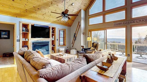 A spacious living room with a wooden ceiling, a fireplace, a large window overlooking Deep Creek Lake, and a television. - 5BR - Chalet - Hot Tub (McHenry)