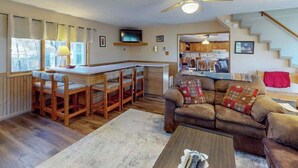 A cozy living room with a sofa, a coffee table, and a TV. There's a kitchen area with a bar and a dining set. - 3BR - Cozy Cabin - Hot Tub (McHenry)