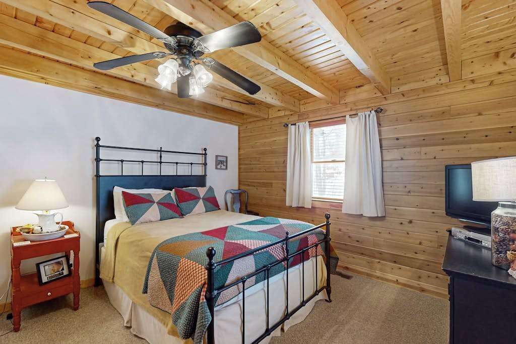 A bedroom with a bed, a ceiling fan, a television, and a window with curtains.