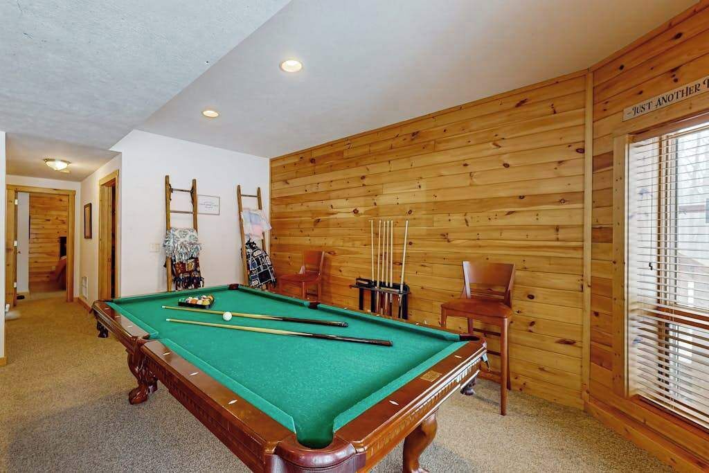 Une table de billard dans une pièce aux murs lambrissés, avec une chaise et une fenêtre équipée de stores.