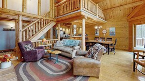 A spacious log cabin interior with a central staircase, a dining area, and a cozy seating arrangement. - Rustic 4BR Cabin - HOT TUB - McHenry (McHenry)