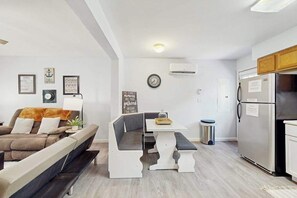 A modern living room with a sofa, a bench, a dining table, and a kitchen area with a refrigerator. - 3BR - Wooded Retreat - Fire Pit (Oakland)