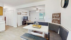 Une cuisine moderne avec des meubles blancs, une table à manger avec un plateau en verre et un coin salon comprenant un banc et un canapé.