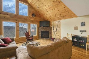 A log cabin interior with a fireplace, wooden floors, large windows, and a cozy seating area.