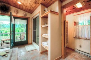 Una habitación con techo de madera, un divisor de vidrio corredizo que da a un espacio exterior, una estantería empotrada y una ventana con cortina blanca.