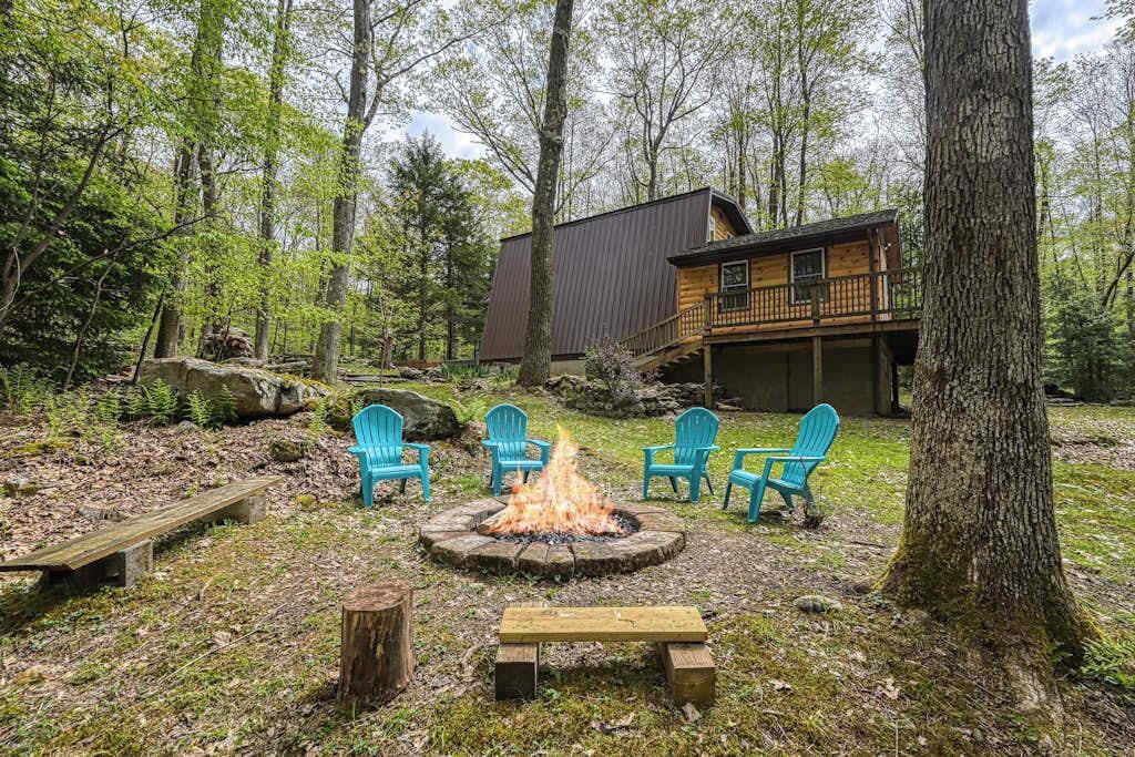 Eine Holzhütte mit Feuerstelle, umgeben von Stühlen, einer Terrasse und einem Wald im Hintergrund.