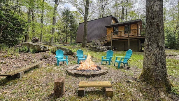 Uma cabana com lareira rodeada por cadeiras, um deck e um cenário de floresta.