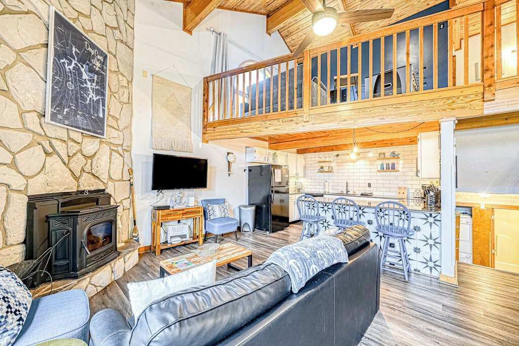 A spacious living area with a stone wall, a fireplace, a television, a balcony with wooden railings, and a kitchen area.