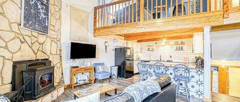 A spacious living area with a stone wall, a fireplace, a television, a balcony with wooden railings, and a kitchen area.