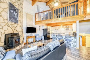 Ein großzügiger Wohnbereich mit Steinwand, Kamin, Fernseher, einem Balkon mit Holzbalkonen und einer Küchenzeile.