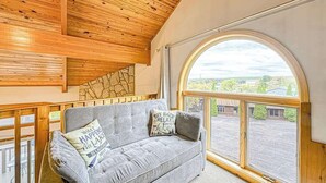 Ein Zimmer mit Holzboden, einem grauen Sofa mit Kissen und einem großen, gewölbten Fenster mit Blick auf Gebäude und Grünflächen.