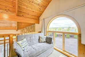 Ein Zimmer mit Holzboden, einem grauen Sofa mit Kissen und einem großen, gewölbten Fenster mit Blick auf Gebäude und Grünflächen.