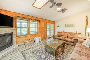 A cozy living room with a fireplace, wooden walls, a ceiling fan, and a large window.