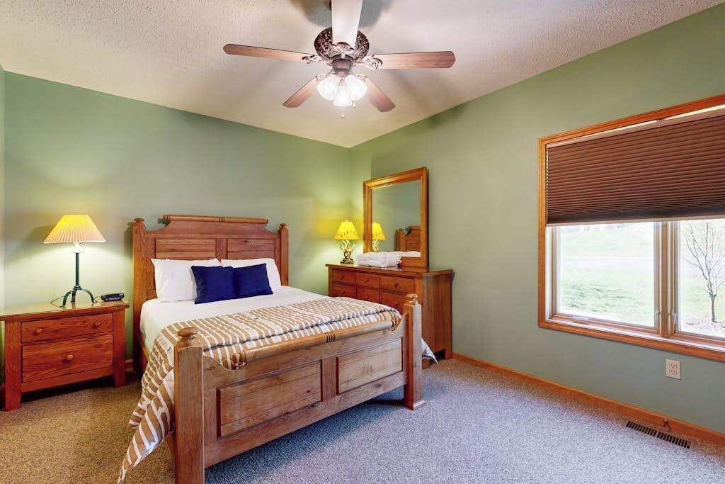 Une chambre à coucher avec un lit en bois, des tables de chevet, un ventilateur de plafond, une fenêtre avec des stores et un miroir.