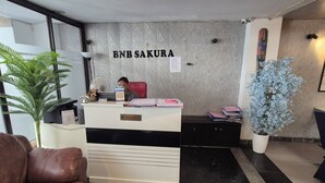 Un mostrador de recepción con un letrero que dice 'BNB SAKURA', una planta en maceta, un sofá de cuero y una mesa de centro con un arreglo floral.