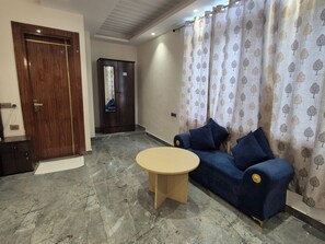 A room with a wooden door, a dark-colored wardrobe, a blue sofa with cushions, a round table, and a patterned curtain. - OnlyStay BnB Golf Course Road (Gurugram)
