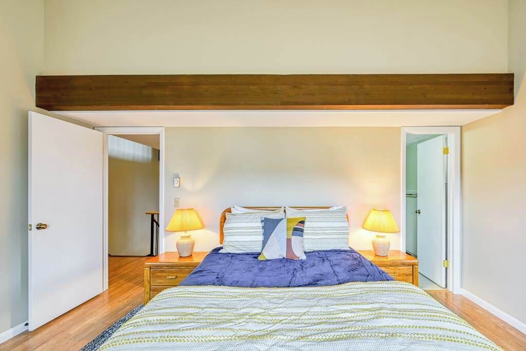 A bedroom with a bed, two bedside tables with lamps, a wooden ceiling beam, and a door leading to another room.