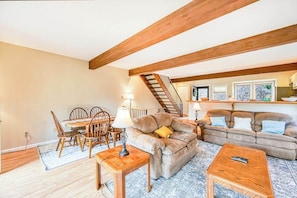 A cozy living room with a dining area, wooden beams, a staircase, and a sofa.
