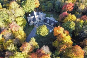 An aerial view of a large, multi-story house surrounded by dense, colorful autumn foliage.
