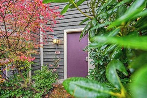 Una construcción de madera con una puerta morada, rodeada de exuberante follaje y un pequeño árbol de hojas rosadas.