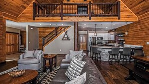 A spacious living area with a high ceiling, a staircase leading to an upper level, and a kitchen area visible through an open door. - Charming 6BR Chalet - HOT TUB - Mountain View (McHenry)