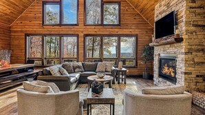 A log cabin interior with a fireplace, large windows, and a cozy seating area. - Charming 6BR Chalet - HOT TUB - Mountain View (McHenry)